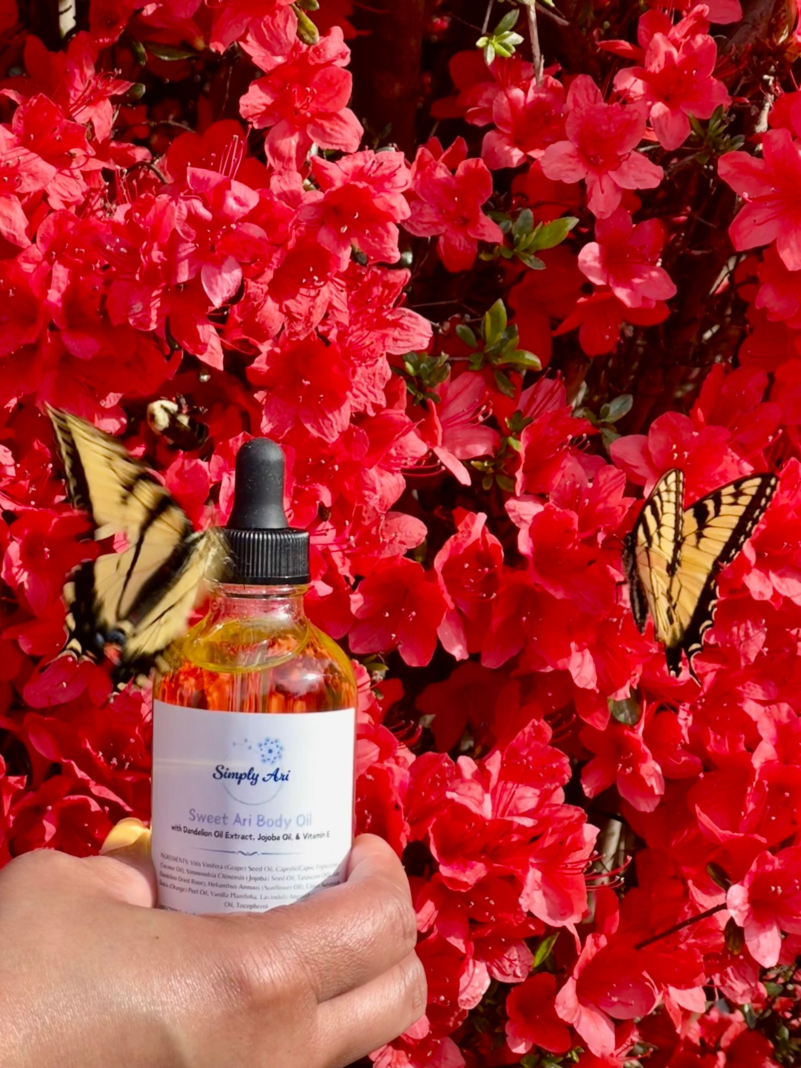 Hand holding Simply Ari Sweet Ari Body Oil against red azaleas with yellow swallowtail butterflies
