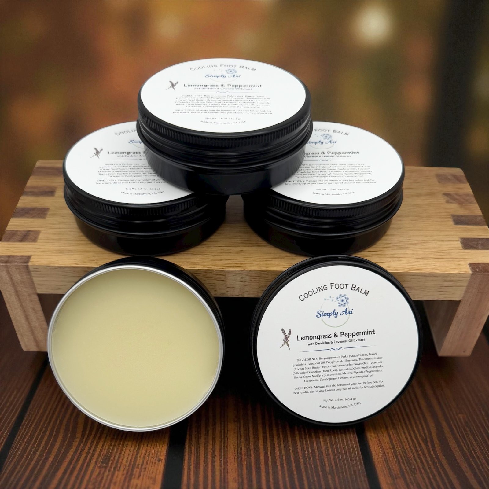 Multiple Simply Ari Cooling Foot Balm tins styled on a wooden riser with one open showing the balm